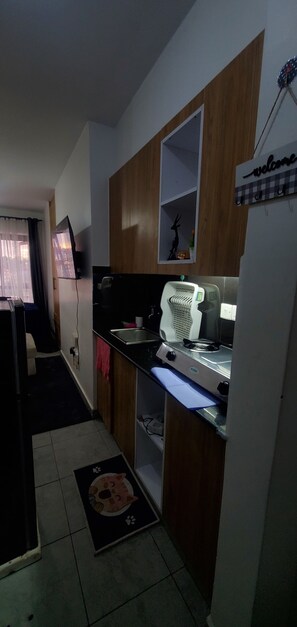 Cozinha compacta com armários de madeira, fogão a gás, pia e uma televisão fixada na parede.