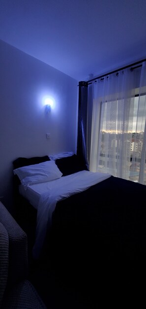 Quarto de hotel com cama, luz noturna, janela com cortinas e vista para prédios.