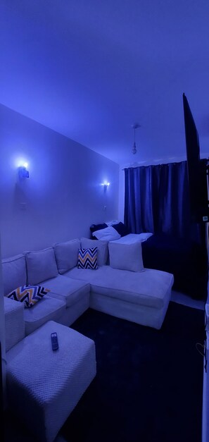 Um quarto com sofá branco em formato de L, um controle remoto no apoio de braço e uma cortina.