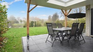 Une terrasse extérieure avec une table et des chaises, un parasol noir et une vue sur une pelouse et des arbres.
