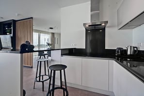 A modern kitchen with white cabinets, a black countertop, and a stainless steel range hood. There are two black bar stools and a potted plant in the background. - Waterfront Apartment Near Beach (Breskens)