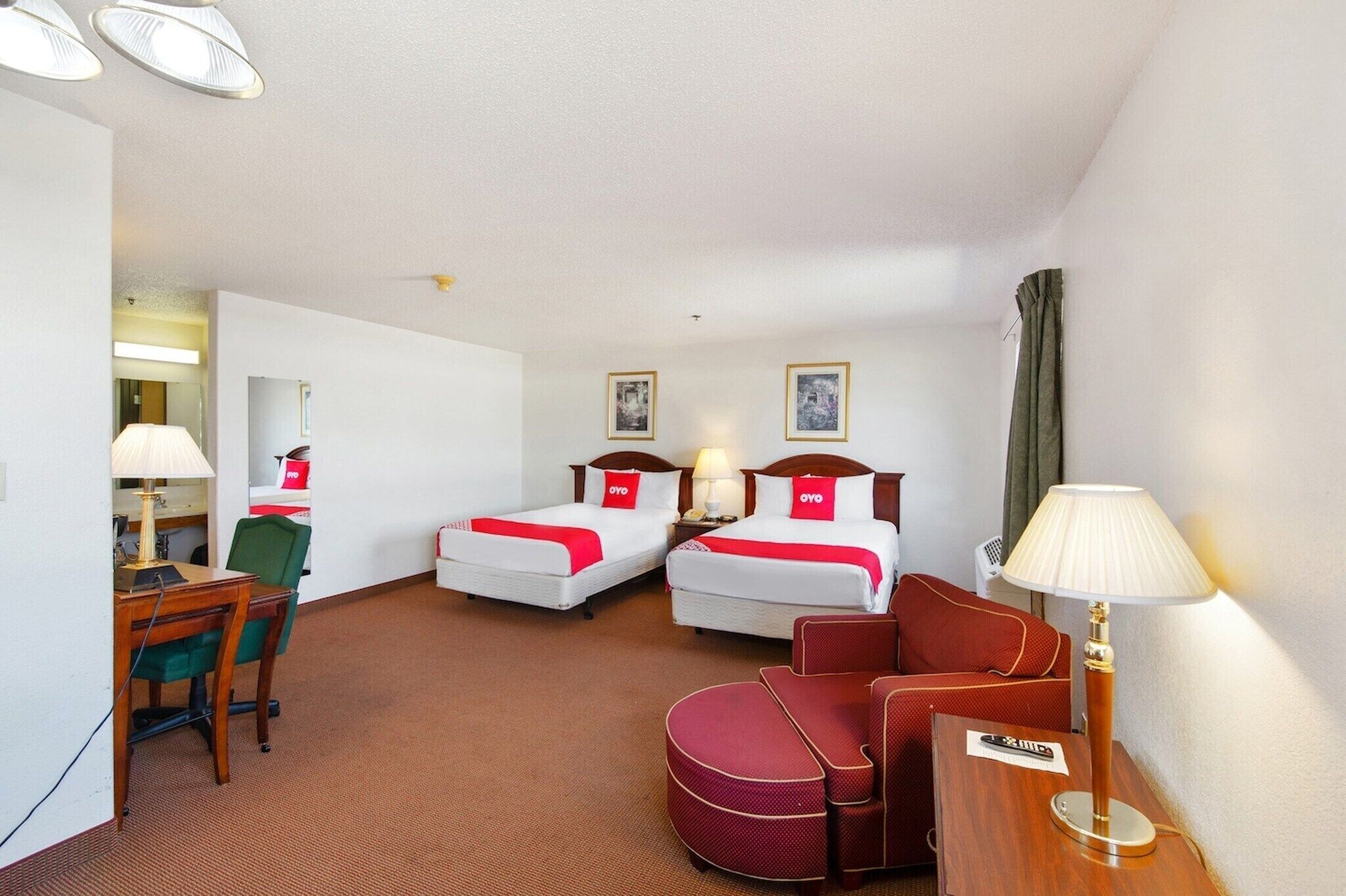 A hotel room with two beds, a desk, a chair, a sofa, and a lamp.