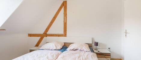 Un dormitorio con techo inclinado, cama con ropa de cama blanca, mesita de noche con lámpara y estructura de vigas de madera.