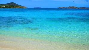 Een strand met helder turquoise water, een zandstrand en heuvels in de verte.