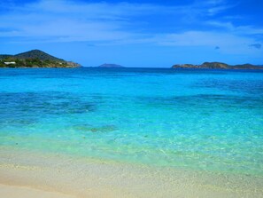 Ein Strand mit klarem türkisblauem Wasser, sandigem Ufer und sanften Hügeln im Hintergrund.
