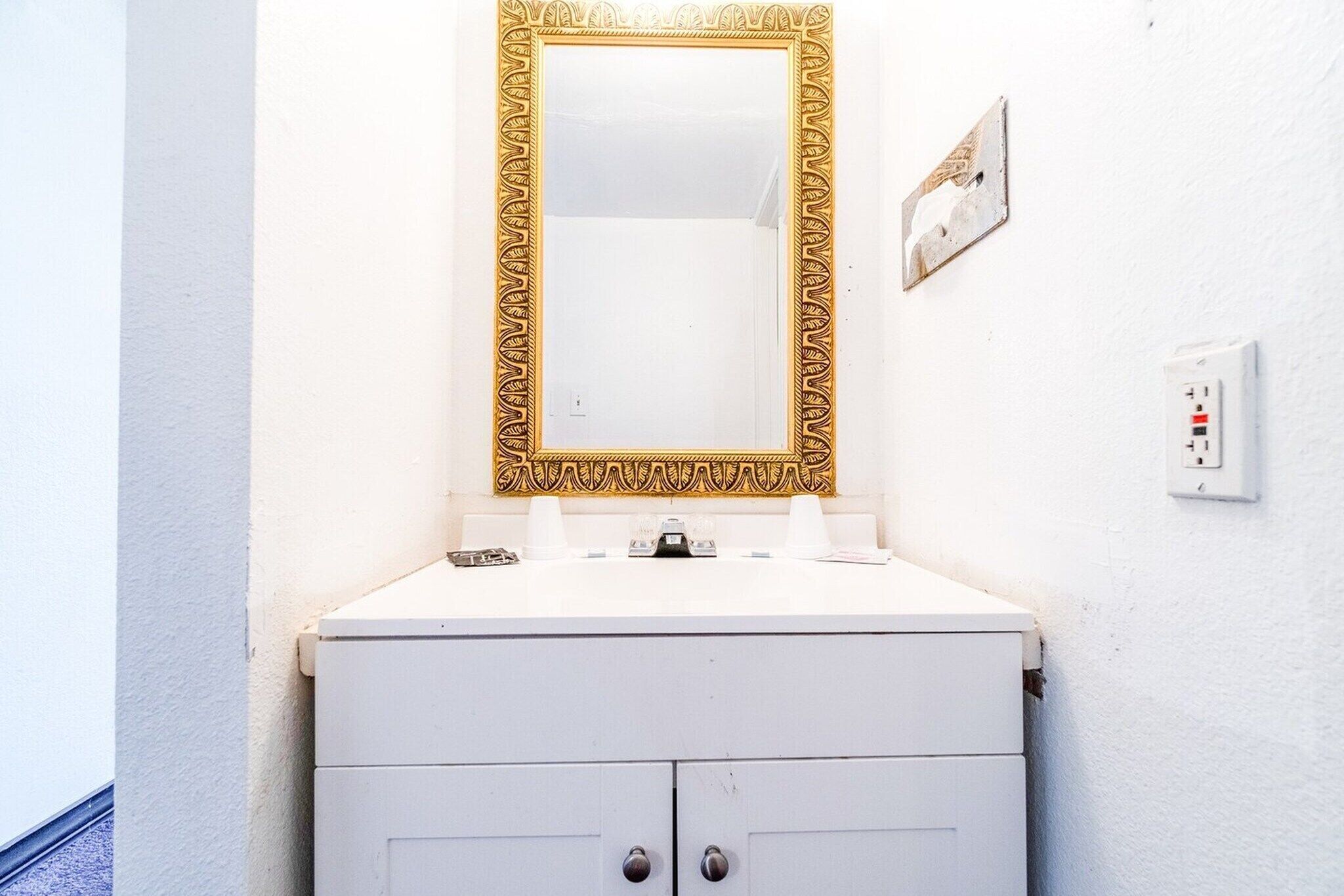 Une salle de bain avec un lavabo blanc, un miroir encadré d’or et un meuble blanc doté de deux tiroirs.