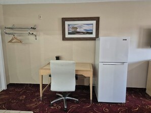 Una habitación pequeña con escritorio, silla, refrigerador, perchero y un cuadro en la pared.