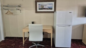 Ein kleines Zimmer mit Schreibtisch, Stuhl, Kühlschrank, Kleiderhaken und einem gerahmten Bild an der Wand.