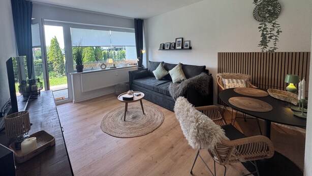 A modern living room with a sofa, coffee table, and a dining area with chairs. There is a large window with a view of greenery outside. - Felicitas Maris - Happiness on the Baltic Sea in Dahme! (Dahme)