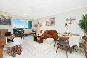 A living room with a sofa, coffee table, dining table, chairs, and a view through a glass door. - Sahara Oasis - Rooftop Pool, CBD & Sunset Balcony (Darwin City)