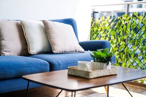 Ein blaues Sofa mit zwei Kissen, ein Couchtisch aus Holz mit einer Pflanze und einer Tasse, und eine grüne, blattbewachsene Wand im Hintergrund.
