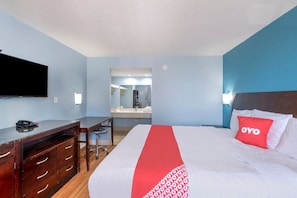 Habitación de hotel con cama, escritorio, televisor de pantalla plana, espejo y baño visible a través de una puerta abierta.