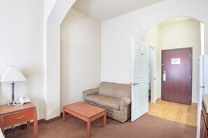 A hotel room with a sofa, a small table, a lamp, a phone, and a door leading to another room. - Premium King Bed with Pull Out (Clute TX)