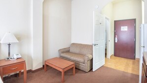 A hotel room with a sofa, a small table, a lamp, a phone, and a door leading to another room. - Premium 2 Queen Bed with Kitchenette (Clute TX)
