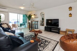 A modern living room with a sofa, coffee table, TV, and a balcony with a view. - Charming | Rooftop Pool, CBD & Sunrise Balcony (Darwin City)
