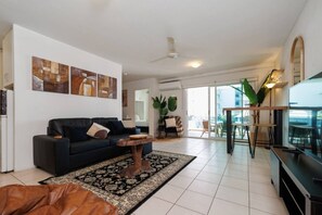A modern living room with a black sofa, a coffee table, and a large TV. There is a balcony with a dining area and a view of the city. - Charming | Rooftop Pool, CBD & Sunrise Balcony (Darwin City)