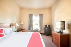 A hotel room with a bed, a TV, a desk with a lamp, a heater, and a window with curtains.
