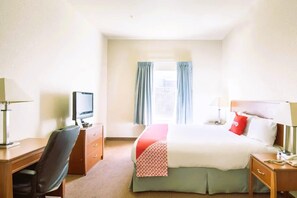 A hotel room with a bed, a desk with a computer, a chair, a bedside table with a lamp, and a window with curtains. - Townhouse Clute Lake Jackson|King Bed (Clute TX)