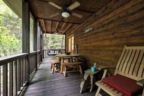 Een houten veranda met een tafel en banken, een plafondventilator en uitzicht op het bos.