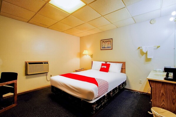 A hotel room with a bed, a desk with a sink, a chair, an air conditioning unit, and a wall-mounted lamp.
