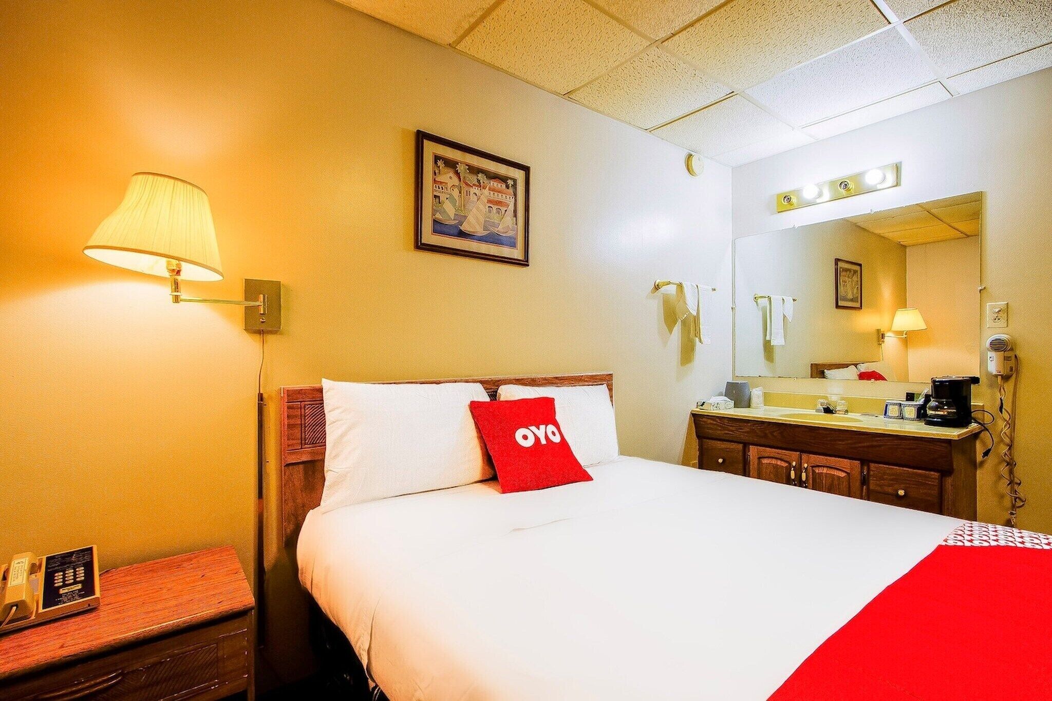 A hotel room with a bed, bedside lamp, a painting on the wall, a mirror above the sink, and a telephone on the nightstand.