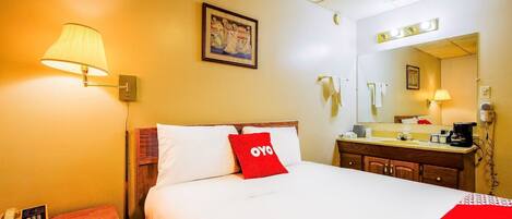 A hotel room with a bed, bedside lamp, a painting on the wall, a mirror above the sink, and a telephone on the nightstand.