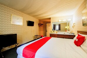 Quarto de hotel com cama, televisão, escrivaninha, espelho e banheiro.