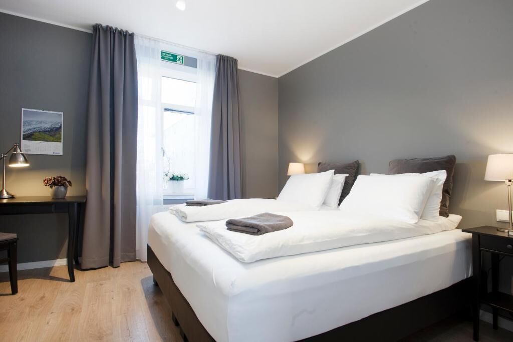 Una habitación de hotel con cama, mesitas de noche, escritorio y ventana con cortinas.
