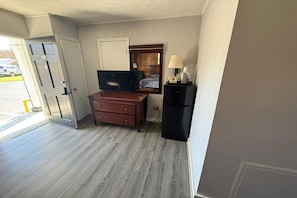 A room with a wooden desk, a television, a small refrigerator, and a door leading outside.