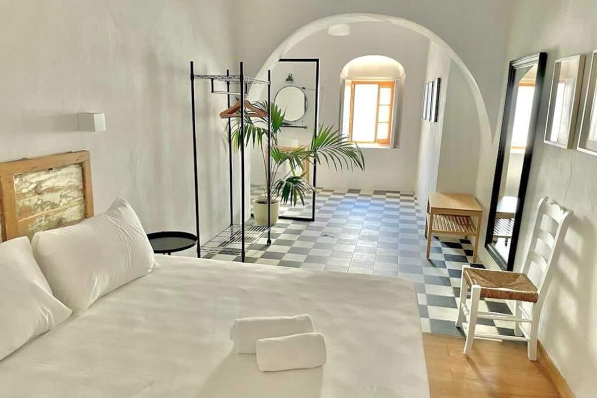 A bedroom with a large bed, a small table, a chair, a potted plant, and a checkered floor.