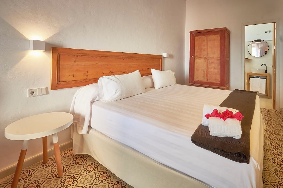 A hotel room with a bed, a small round table, a wooden headboard, a wardrobe, a mirror, and a bathroom with a sink and towel rack.