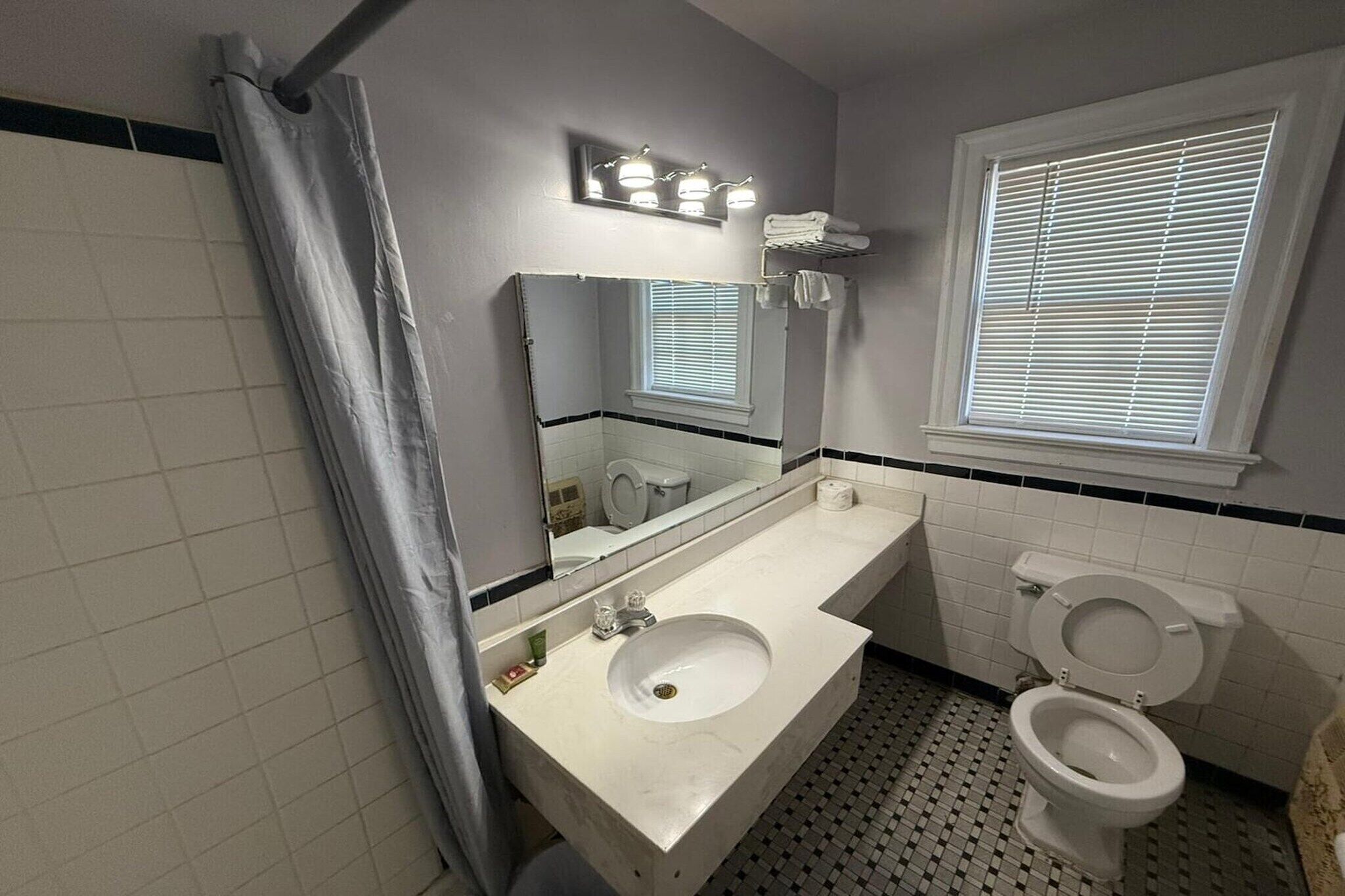 Une salle de bain avec un lavabo blanc, un miroir, des toilettes et une fenêtre avec des stores.