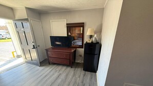 Une pièce comprenant un bureau en bois, une télévision, un petit réfrigérateur et une porte donnant sur l’extérieur.