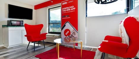 A modern hotel room with a red accent wall, a flat-screen TV, a microwave, a glass table, and two red chairs.