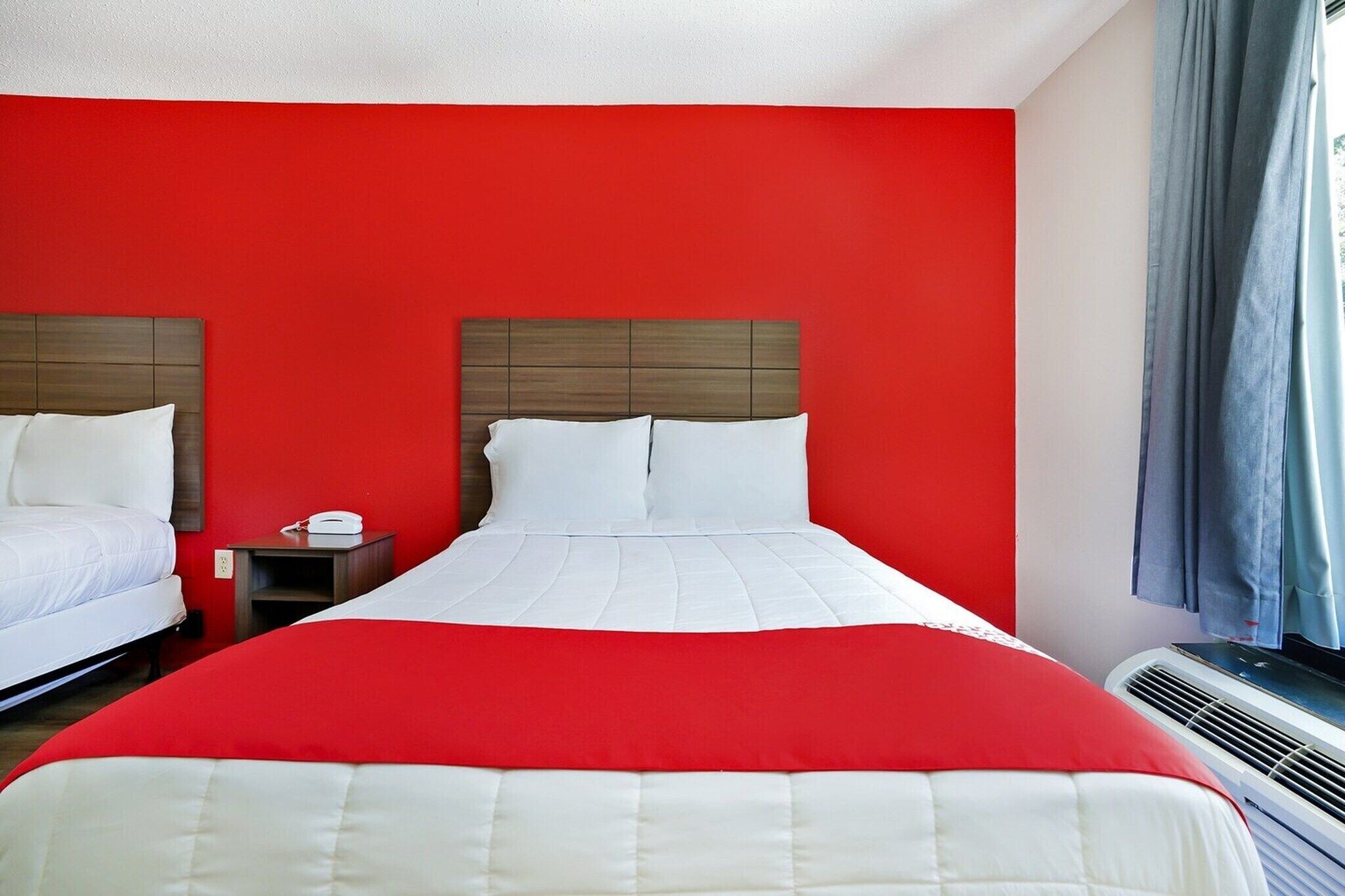 A hotel room with two beds, a red wall, a white bedspread, a wooden headboard, a bedside table with a lamp, and a window with curtains.