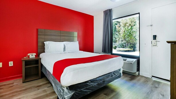 Une chambre d’hôtel avec un mur rouge, un lit avec une literie blanche, une table de chevet avec un téléphone et une fenêtre donnant sur des arbres.
