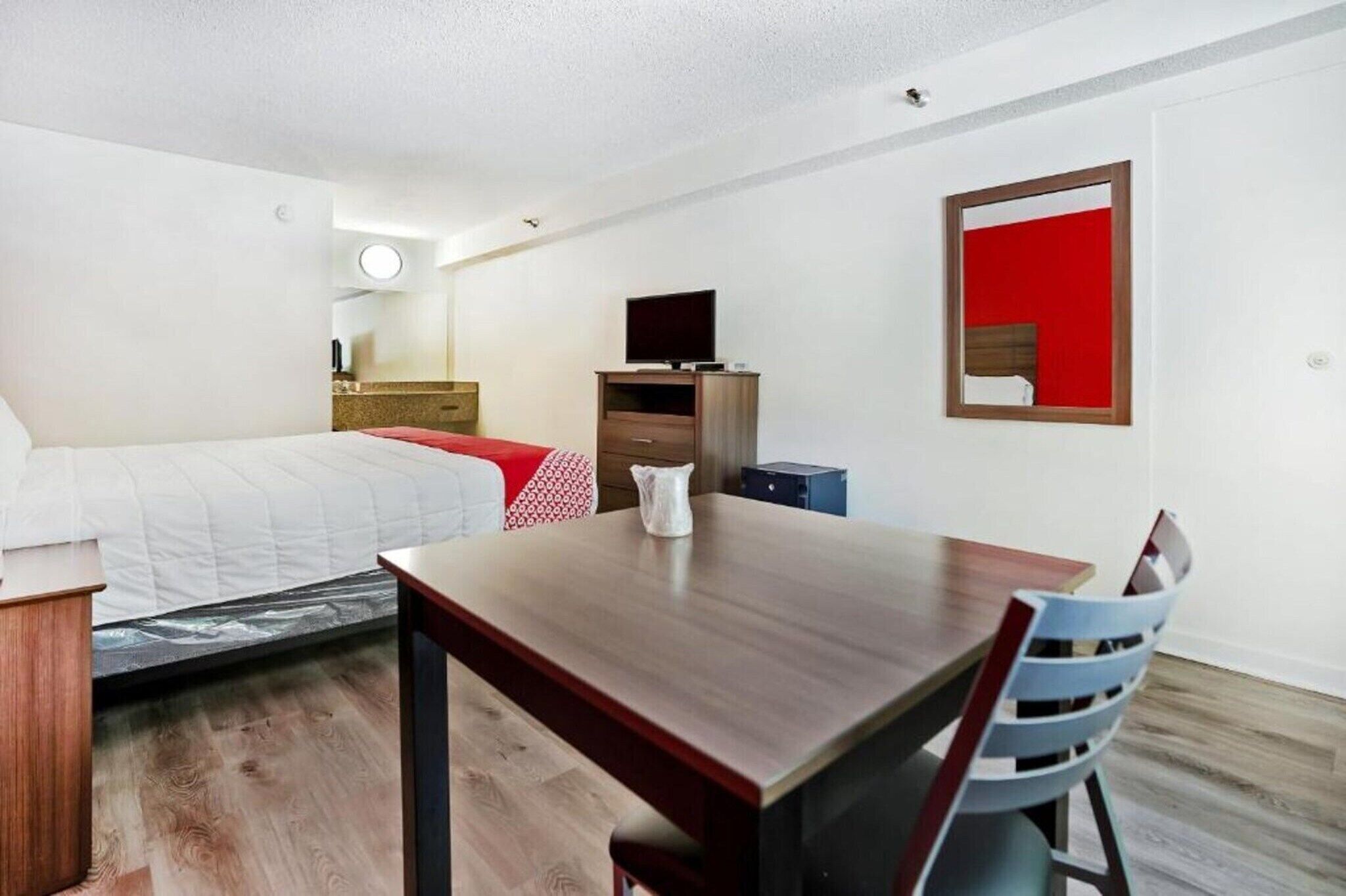 A hotel room with a bed, a wooden table, a chair, a TV, a dresser, and a mirror.