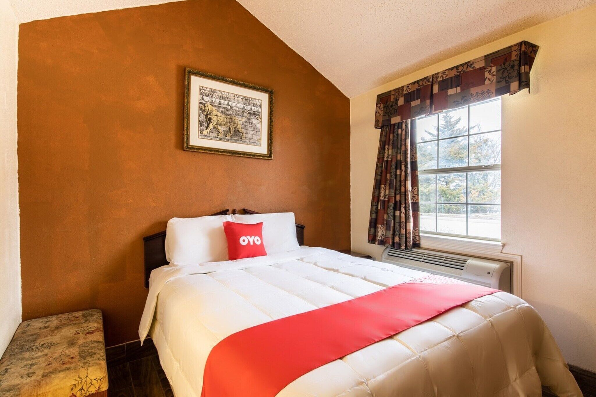 Een hotelkamer met een bed, een raam met gordijnen, een aan de muur hangend schilderij en een bankje.