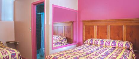 A bedroom with a pink wall, a wooden headboard, a bed with a patterned bedspread, a mirror, and a door leading to a bathroom.