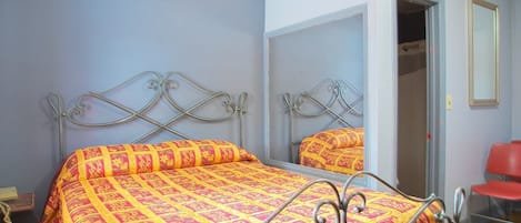 A bedroom with a metal bed frame, a patterned bedspread, a mirror, and a red chair.