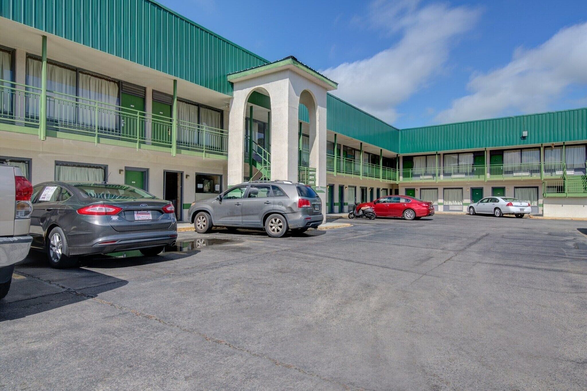A motel with a green facade, multiple parking spaces, and a central entrance with a small archway.