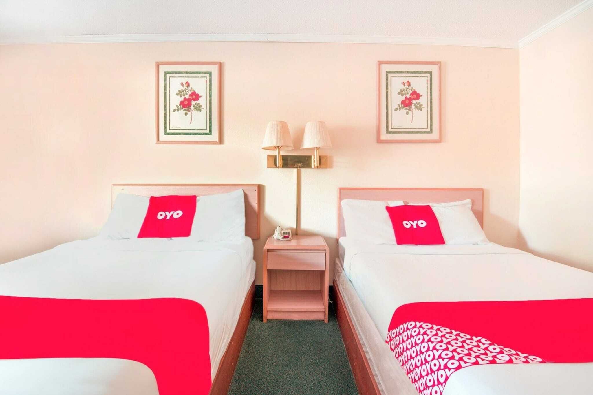 A hotel room with two beds, each with a red OYO branded pillow, a nightstand with a lamp, and two framed floral pictures on the wall.