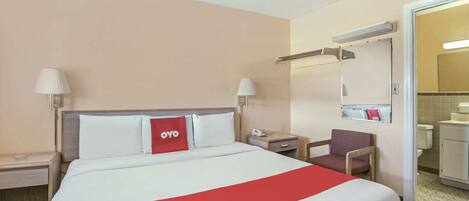 A hotel room with a bed, bedside tables, a chair, a mirror, and a bathroom visible through an open door.