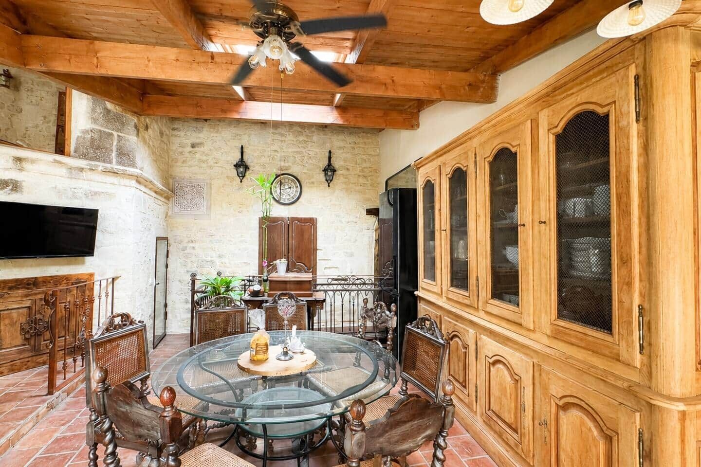 Una stanza con un tavolo rotondo in vetro, sedie in legno, una ventola a soffitto e un'ampia credenza in legno con ante in vetro.
