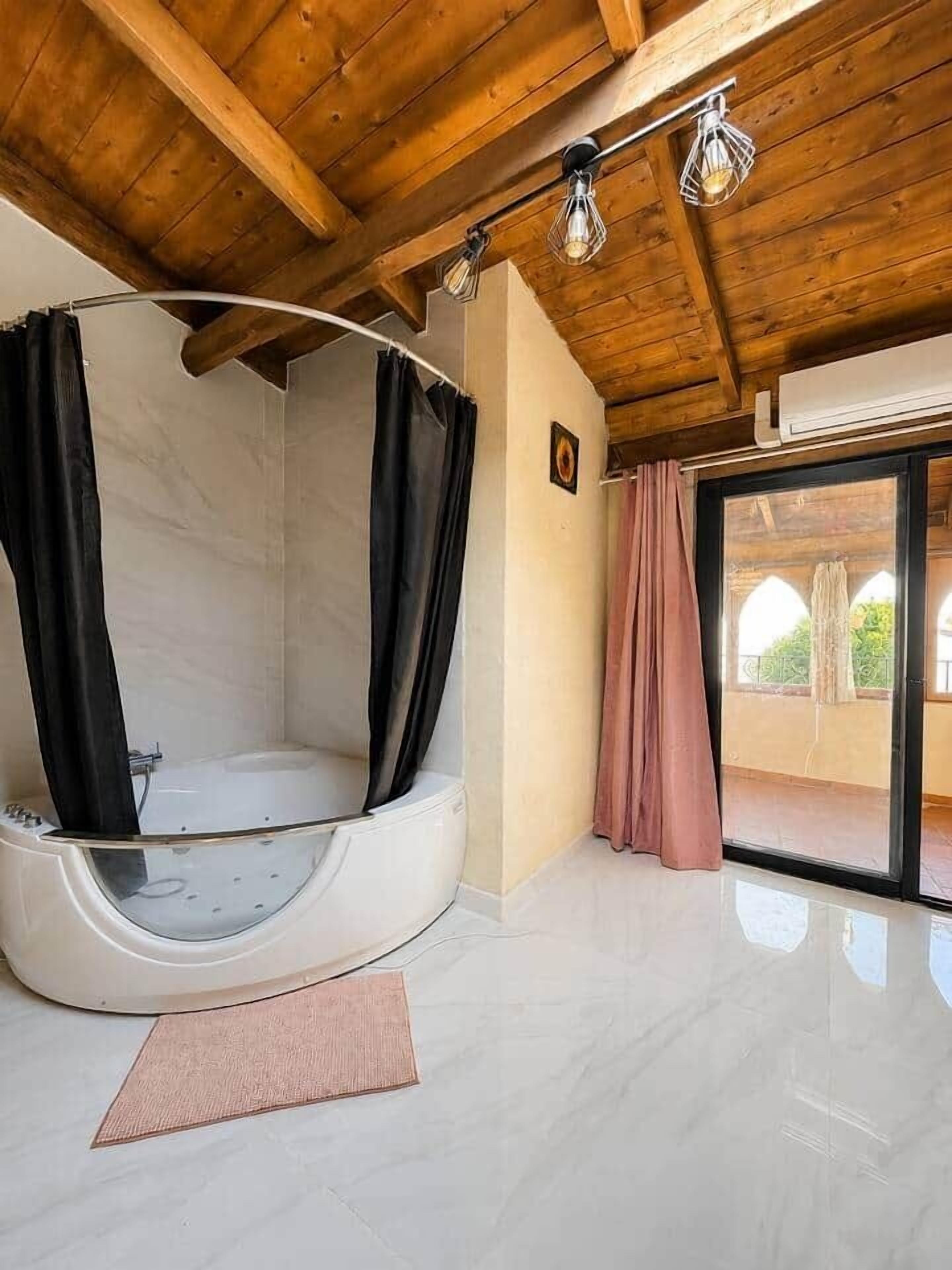 A modern bathroom with a freestanding bathtub, wooden ceiling beams, and a view through a glass door.