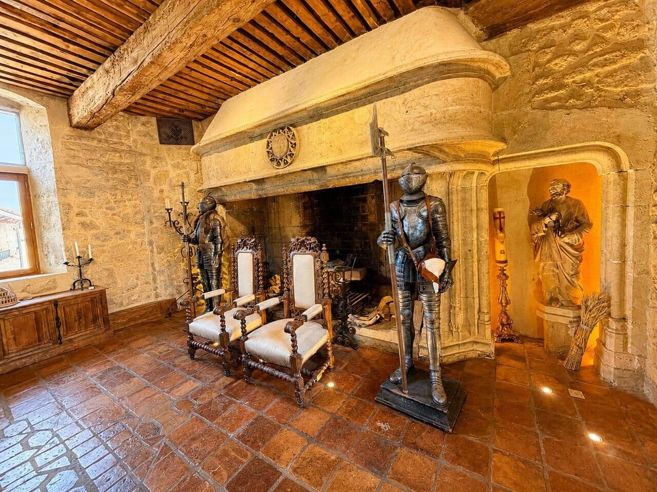 A stone-walled room with a fireplace, wooden chairs, and a knight statue.
