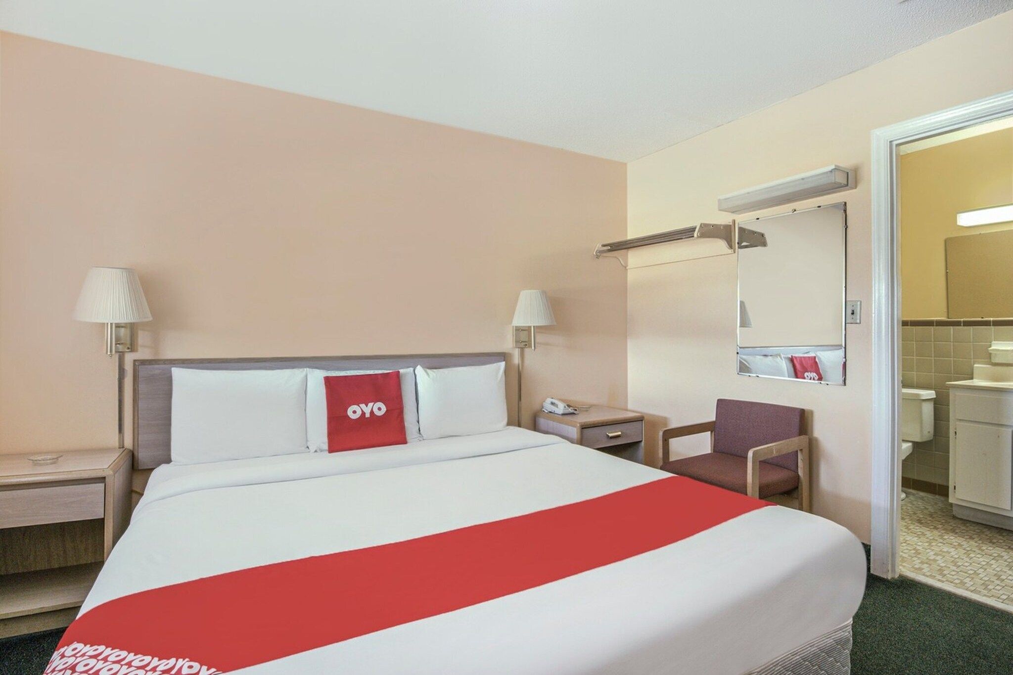 A hotel room with a bed, two bedside tables, a chair, a mirror, and a bathroom visible through an open door.