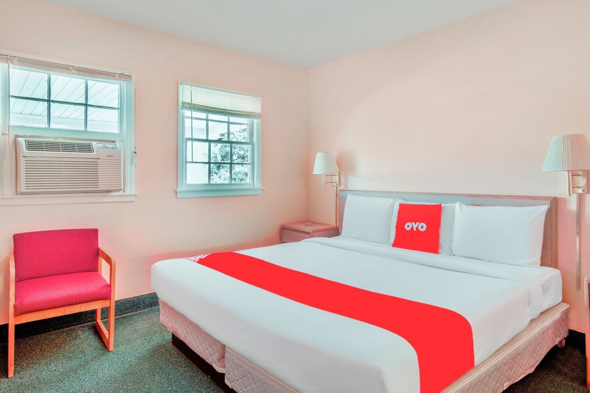 Une chambre d’hôtel avec un lit, une chaise, une table de chevet, une lampe et deux fenêtres.