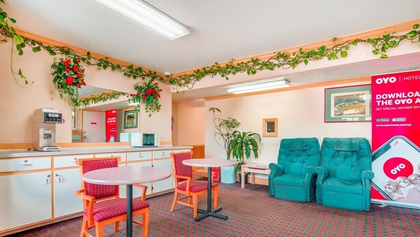 A hotel lobby with a reception desk, two round tables with chairs, a large potted plant, and teal armchairs.