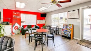 A hotel room with a red accent wall, a ceiling fan, a dining area with a round table and chairs, a sofa, a TV, and a window with blinds.